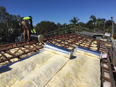 Worker installing reflective foil-backed roof insulation between wooden trusses under clear blue sky.